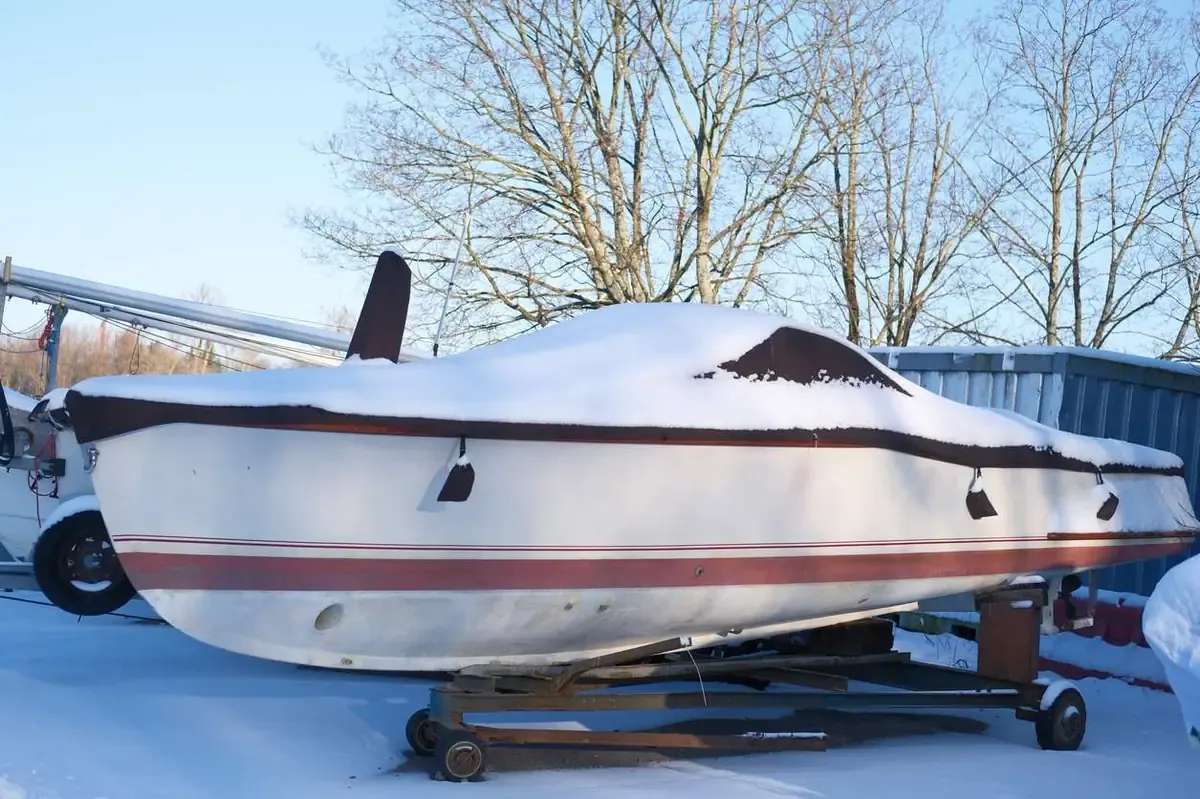 A small boat covered in snow while stored outdoors on a trailer, with bare winter trees and bright sunlight.